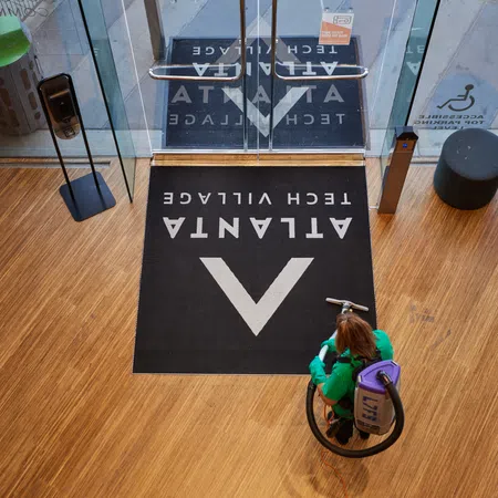 Woman cleaning hardwood floor near glass doors with Atlanta Tech Village logo mat in front entrance.
