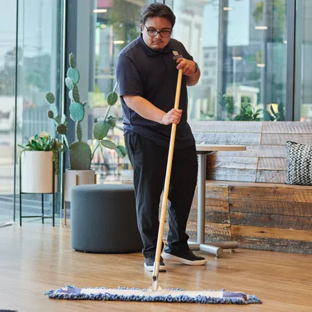 Man mopping wooden floor in bright modern cafe with indoor plants and large glass windows during daytime.