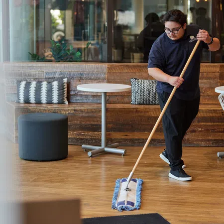 Man mopping a clean wooden floor in a modern cafe with tables and chairs in the background