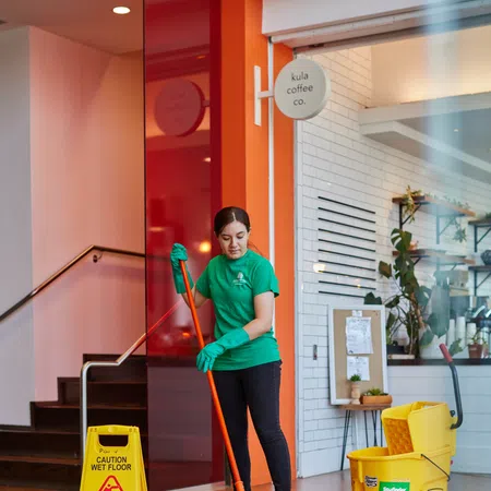 Worker mopping floor near caution wet floor sign inside a coffee shop with wooden flooring and cleaning equipment.
