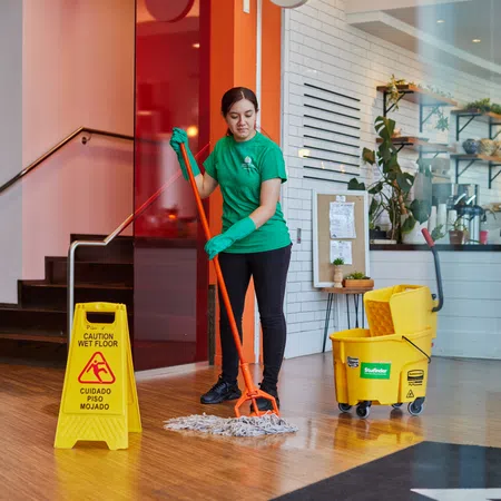 Janitor in green shirt mops wooden floor near caution wet floor sign and yellow cleaning bucket in bright lobby.