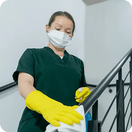 Woman in scrubs and yellow gloves disinfecting stair railing with spray and cloth indoors.