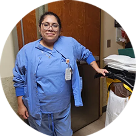 Smiling healthcare worker in blue scrubs and glasses standing in a hospital room next to medical supplies.