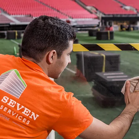 Worker in orange shirt cleaning glass barrier inside a large empty stadium with red seats and green field.