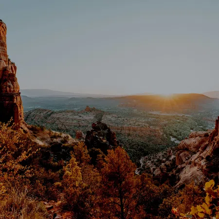 Sunrise over rocky red cliffs and autumn trees in a vast desert landscape with distant mountains.