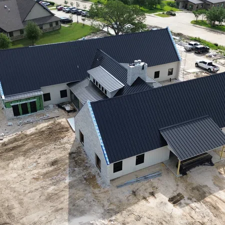 Aerial view of a new commercial standing seam metal roof installation on a dental office under construction in College Station, Texas, featuring dark charcoal metal roofing across multiple gabled sections with a stone chimney and surrounding medical office buildings.