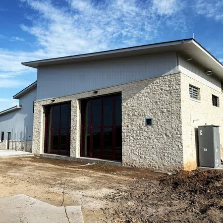 Exterior view of a new commercial building under construction featuring a light limestone stone facade installed by Mattco, with metal panel accents, tall red-framed glass windows, and a standing seam metal roof under a clear Texas sky.