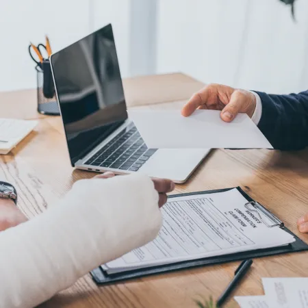 Person with arm cast handing an insurance claim form to a professional at office desk with laptop.