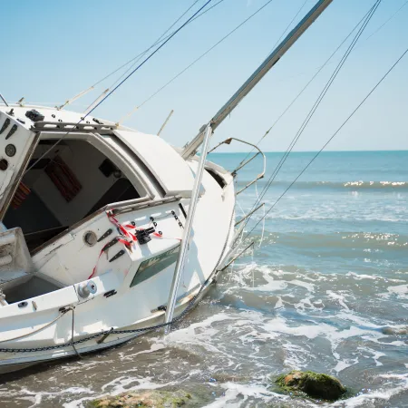 White sailboat washed ashore on rocky beach under clear blue sky with calm ocean waves in the background