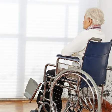 Elderly woman sitting in a wheelchair facing a bright window in a quiet room.