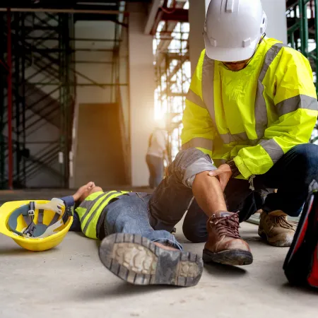 Construction worker providing first aid to injured colleague lying on concrete floor at site.