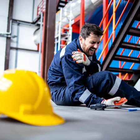 Injured worker in blue safety gear sitting on warehouse floor holding his arm in pain near a yellow hard hat.