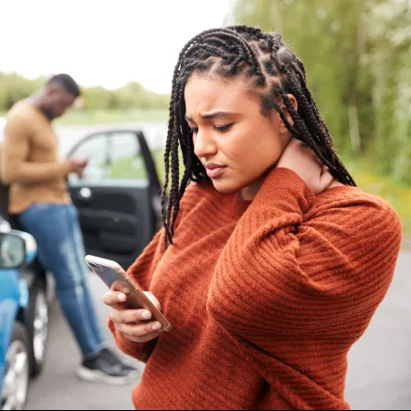 Worried woman checking phone after a car accident, with a man using his phone near damaged vehicles on a road.