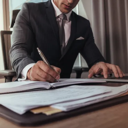 Businessman in suit writing on documents while using a laptop in an office setting with natural light.