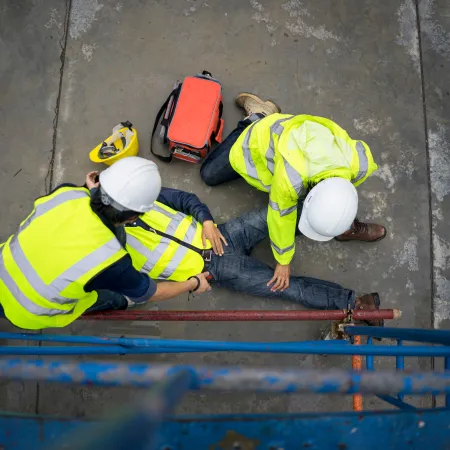 Two construction workers help an injured colleague lying on the concrete floor at a worksite wearing safety gear.