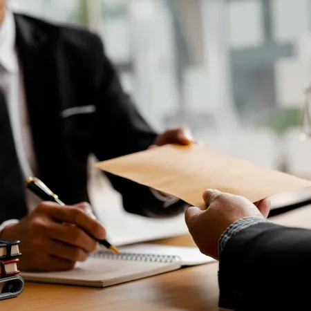 Two business professionals exchanging a brown envelope during a meeting at an office desk with legal books and scales.