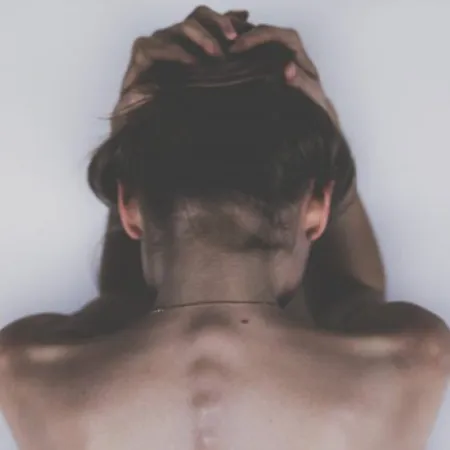Person seen from behind with hands holding the head, emphasizing back spine and shoulder muscles.