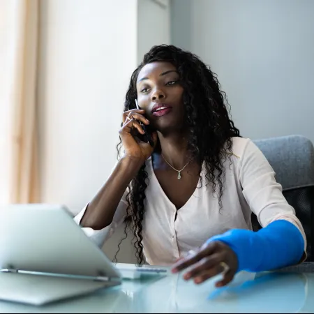 Young woman with blue arm cast talking on phone while working on tablet in bright office space