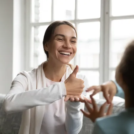 Smiling woman communicating with child using sign language in a bright room with large windows.