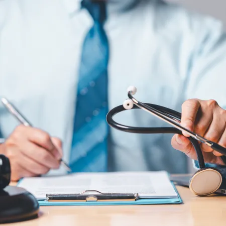 Person in dress shirt and tie holding stethoscope and writing on clipboard next to laptop and gavel