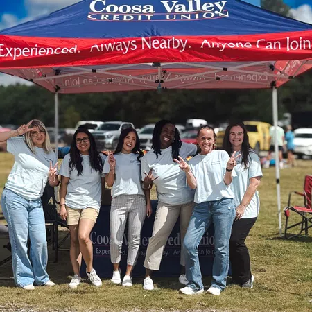 Group of six women posing and smiling under a Coosa Valley Credit Union event tent outdoors on a sunny day.