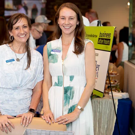 Two smiling women standing at a business networking event, dressed in white with name tags, indoor setting.