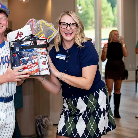 Smiling baseball player in uniform receiving a gift basket from a woman in glasses inside a bright room.
