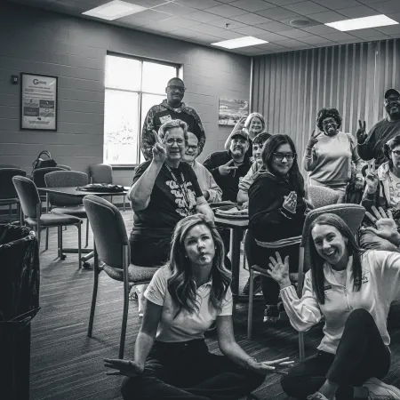 Black and white group photo of diverse people in a room smiling and making hand gestures, seated and standing.