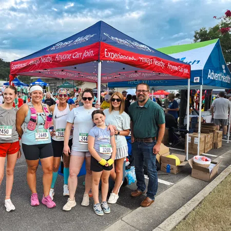 Group of runners with race bibs posing under event tents at a community outdoor race event on a cloudy day.