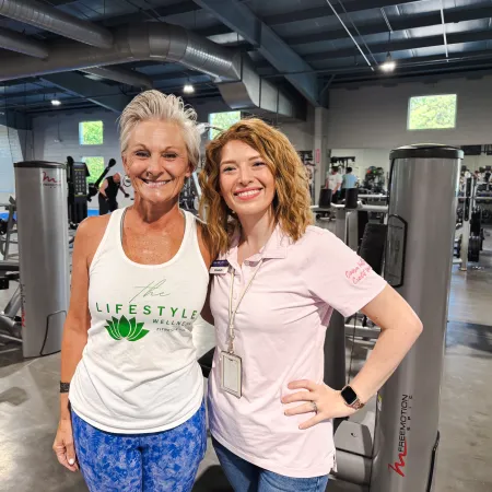Two smiling women posing together in a bright gym filled with fitness equipment and weights.
