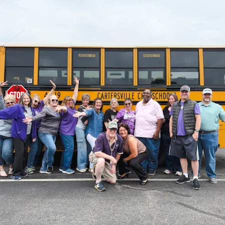 Group of smiling adults posing happily in front of a yellow Cartersville City Schools bus in a parking lot.