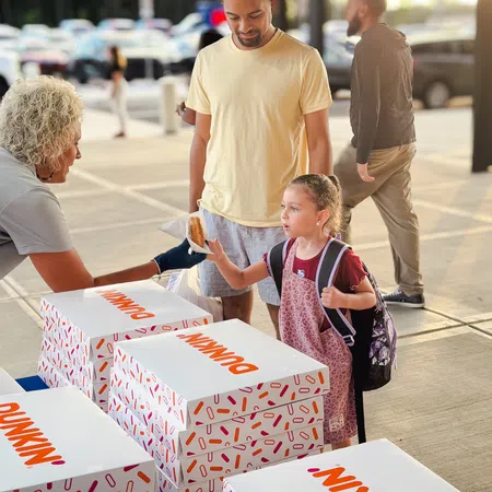 Young girl with backpack receives a Dunkin' donut from a vendor at an outdoor event with her father nearby.