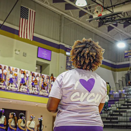 Basketball gym with cheerleaders and a woman wearing an I love CVCU shirt facing the court under an American flag.
