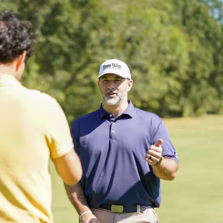 Two men on golf course, one in yellow shirt with back to camera, the other in navy shirt and white cap speaking.