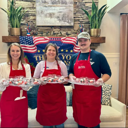 Three people wearing red aprons holding trays of cupcakes in front of a patriotic banner and stone wall.
