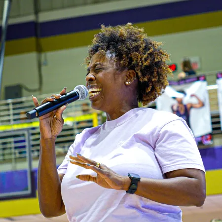 Woman happily speaking into a microphone inside a gymnasium with basketball scoreboard and banners in the background