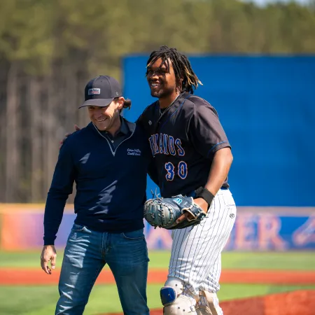 Two men smiling and embracing on a sunny baseball field with one in uniform and the other in casual wear.