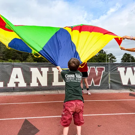 Children and adults playing with a colorful parachute on a running track in an outdoor sports area.