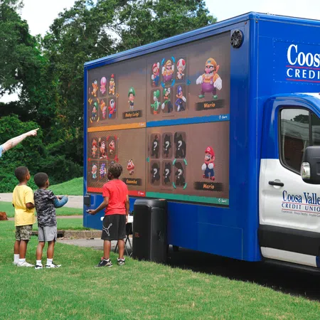 Children and adult engage with a Coosa Valley Credit Union gaming truck featuring a large Mario Kart video game screen outdoors.