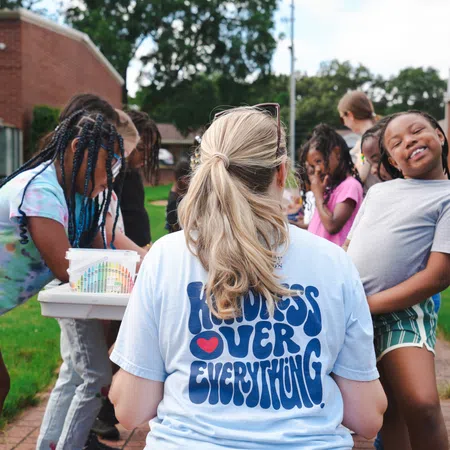 Woman with kindness over everything shirt interacts with smiling children outside near red brick buildings on a sunny day