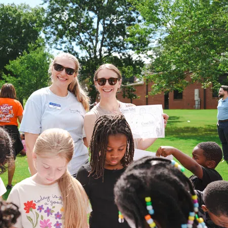 Two smiling women with sunglasses and name tags stand behind a group of children engaged in an outdoor activity on grass.