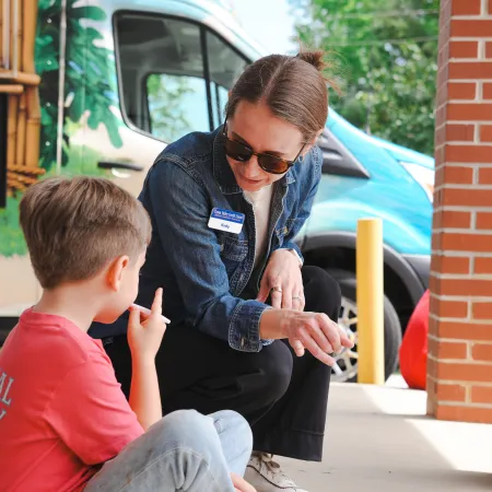 Woman in sunglasses and denim jacket talking to young boy in red shirt near a colorful van outdoors.
