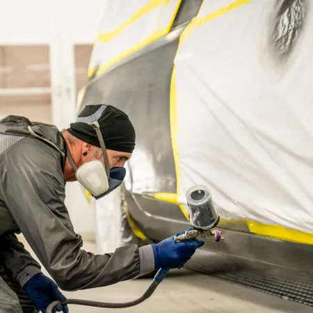 Worker in protective gear spray painting a car body in a professional auto repair shop with masking tape applied.