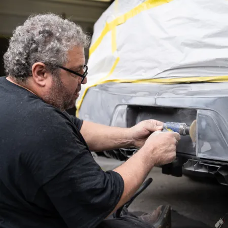 Man sanding a car bumper in a workshop with protective masking tape applied on the car body.