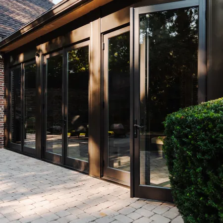 Tall glass patio doors with dark frames opening to a paved outdoor area beside green shrubbery and brick wall.