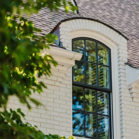 Close-up of an arched black Infinity by Marvin window on a white brick house with a brown shingle roof and greenery.