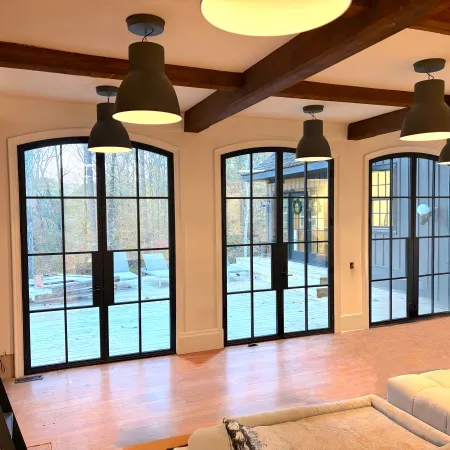 Bright living room with large black-framed glass doors, exposed wooden beams, pendant lights, and a light sofa.