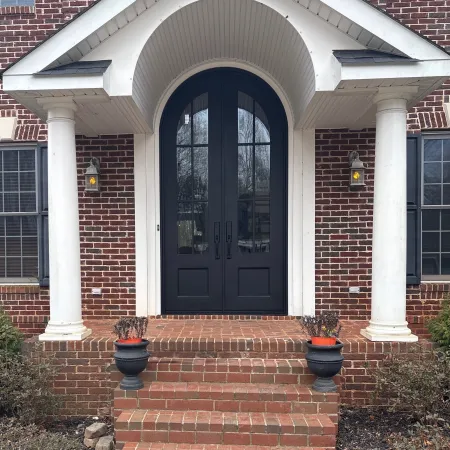 Brick house entrance with a black iron arched double doors, white columns, and brick steps with planter pots.