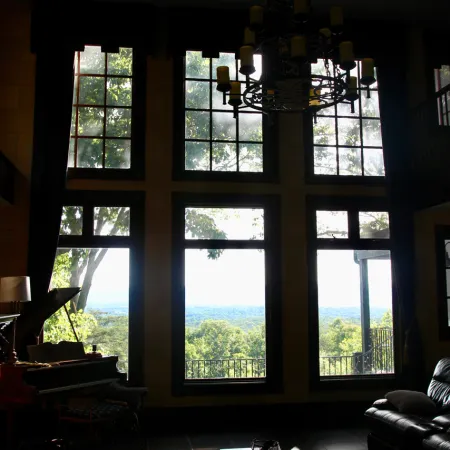 Sunlight filters through large windows illuminating a cozy living room with a chandelier, piano, and leather furniture.
