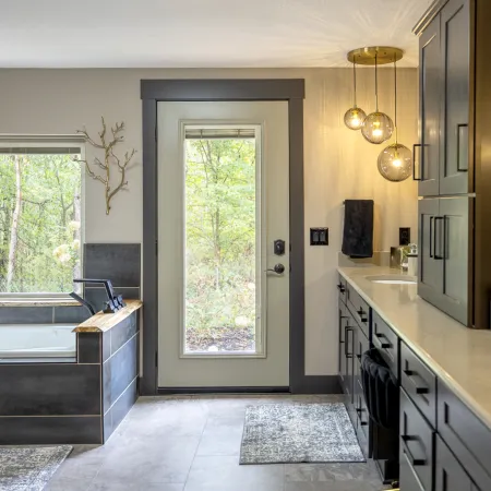 Modern bathroom with dark tile tub, glass door to outdoors, double vanity, and hanging globe lights.